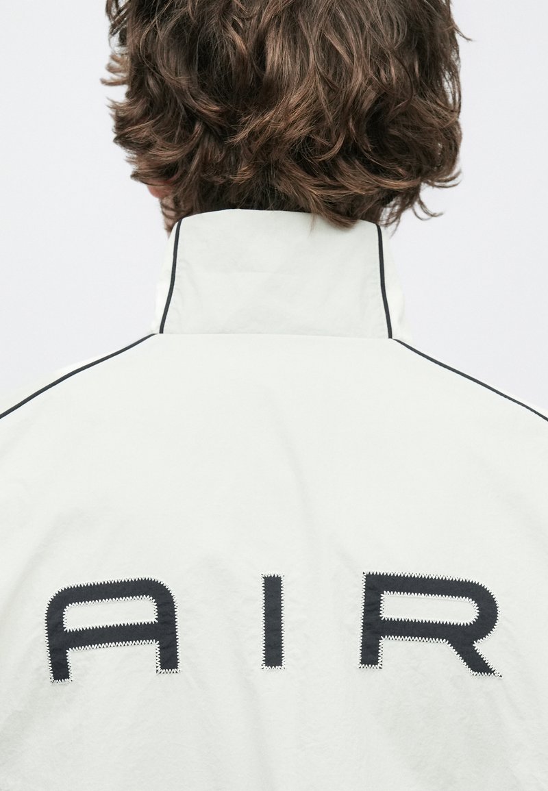 Chaqueta blanca con cuello alto, detalles de costuras negras y grandes letras que forman la palabra "AIR" en la espalda. Material liso y ligero.