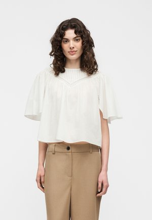 Γυναίκα με σγουρά σκούρα μαλλιά φοράει φαρδιά λευκή μπλούζα με φαρδιά μανίκια και μπεζ ψηλόμεσο παντελόνι, στέκεται μπροστά σε απλό φόντο.