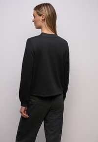 Musta collegepaita, jossa on sileä pinta ja pyöreä pääntie, pitkät hihat ja hieman lyhyt mitoitus. Yhdistetty tummanharmaisiin housuihin.