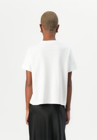 T-shirt blanc à manches courtes avec une coupe droite et une texture douce. La vue arrière montre un design simple et une coupe décontractée, associé à une jupe noire.