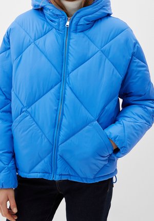 Blue quilted puffer jacket with a front zipper and hood, worn over a white turtleneck, paired with dark pants. One hand in pocket.