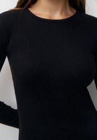 Haut noir à manches longues côtelé avec un col rond. La texture du tissu semble lisse et ajustée, mettant en valeur la silhouette sans ornements.