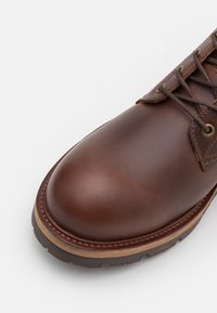 Hnědá kožená bota s kulatou špičkou, pevnou gumovou podešví a kontrastním béžovým prošitím podél okraje a dírek na tkaničky.