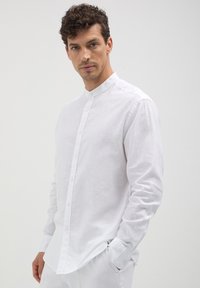 Mann mit lockigem Haar, der ein enganliegendes weißes Hemd mit Mandarin-Kragen und Knöpfen sowie weiße Hosen trägt, steht vor einem schlichten hellen Hintergrund.