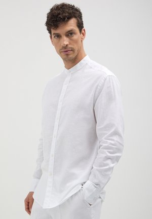 Mann mit lockigem Haar, der ein enganliegendes weißes Hemd mit Mandarin-Kragen und Knöpfen sowie weiße Hosen trägt, steht vor einem schlichten hellen Hintergrund.