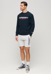 Marineblå sweatshirt med "SUPERDRY SPORTSWEAR" logo, parret med lysegrå shorts med marineblå tekst. Nedarvet fodtøj fuldender outfittet.