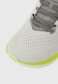 Jasnoszary but sportowy z oddychającą cholewką z siateczki, ciemnoszare sznurowadła i neonowozielona krawędź podeszwy, widziany z kąta od góry.