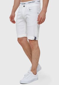 Shorts en jean blanc avec des poignets retournés, présentant un bouton de fermeture et une taille avec le logo "Calvin Klein". Portés avec des baskets blanches.