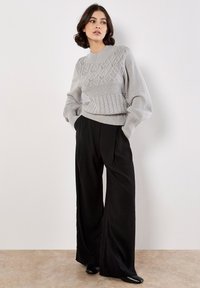Grauer Strickpullover mit strukturiertem Rautenmuster, hohem Kragen und weiten Ärmeln, kombiniert mit schwarzen weiten Hosen und schwarzen Schuhen.
