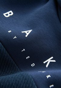 Námorná tkanina so štruktúrovaným pletením a hladkými časťami. Odvážne biele písmená tvoria "BAK" s menším textom "by TED". Jasný kontrast materiálov.