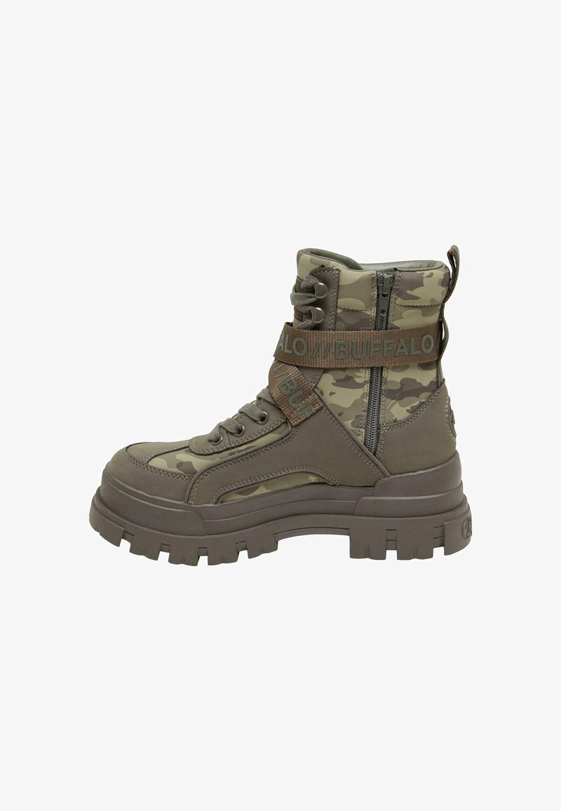 Grüne Ankle-Boots mit Camouflage-Muster, einer klobigen Sohle, einem Reißverschluss und einem Stoffriemen, der den Text "BUFFALO" trägt. Das Material scheint synthetisch und strukturiert zu sein.