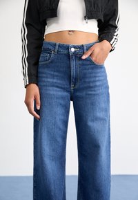 Jean en denim bleu moyen à taille haute et jambes larges, avec surpiqûres jaunes, cinq poches et fermeture par bouton. Texture lisse.