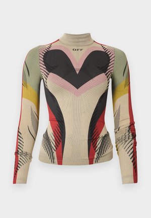 Nő, aki színes, testhez simuló, geometriai mintás overallt visel hosszú ujjakkal és fekete, magas sarkú szandált, egy egyszínű háttér előtt állva.