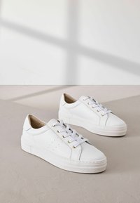 Des baskets blanches en cuir texturé, avec des bouts ronds, des semelles blanches plates et des œillets dorés pour les lacets. Design minimaliste avec des détails perforés.