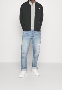 Svart zip-up jacka över en grå skjorta med krage, kombinerat med ljusblå slitna jeans och vita sneakers.