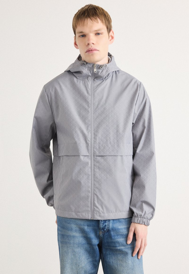 Šviesiai pilkas zip-up striukė su kapišonu, su geometriniu tekstūros raštu, elastingomis rankogaliais ir tiesiu siluetu. Sujungta su mėlynomis džinsomis.