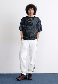 Sort grafisk t-shirt med stort print, hvide træningsbukser og sorte sneakers med røde detaljer. Model står foran en neutral baggrund.