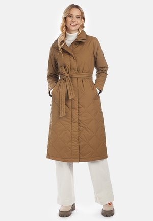 Vrouw draagt een lange gewatteerde bruine jas met riem over witte broek en beige laarzen, glimlachend, staand met handen in de zakken.