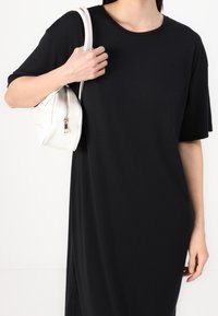 Musta ylisuurikokoinen t-paidamekko, jossa on lyhyet hihat. Malli pitää kädessään pientä valkoista käsilaukkua, jossa on vetoketjullinen suljenta ja pyöreä muoto.