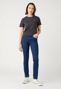 Camiseta de manga corta gris oscuro con cuello redondo. Llevada con jeans ajustados azules y zapatillas blancas de caña alta. Diseño simple, sin patrones notables.