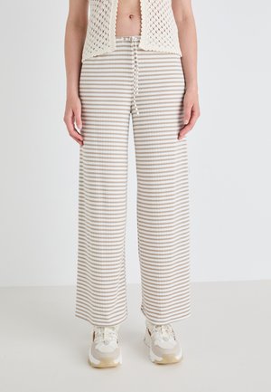 Pantalons larges rayés en beige et blanc cassé, avec une taille à cordon et un tissu doux et texturé. Associés à des baskets chunky.
