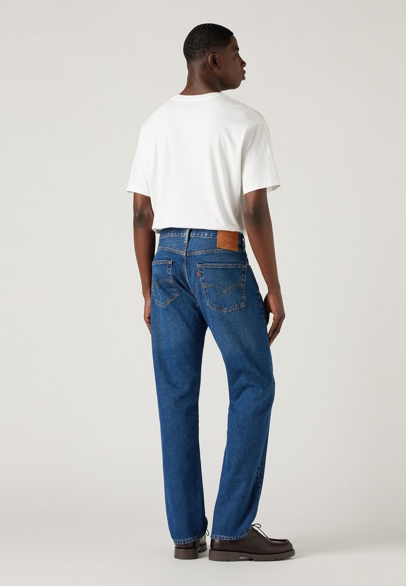 Enges weißes T-Shirt, kombiniert mit blauen Jeans aus Denim, die ein Lederpatch, Gesäßtaschen und einen geraden Schnitt aufweisen. Braune Schuhe runden den Look ab.