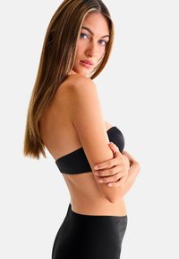Femme aux cheveux longs et raides portant un haut noir sans bretelles et un bas noir taille haute, regardant par-dessus son épaule avec les bras croisés.