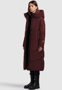 Burgundy long puffer coat with a high collar, zip closure, and quilted pattern. Paired with black ankle boots. Smooth texture.
