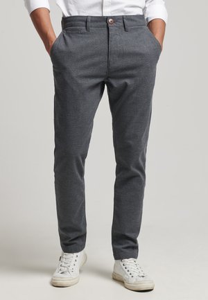 Mężczyzna w szarych, dopasowanych spodniach i białych trampkach, z rękami w kieszeniach, stoi na jasnym, jednolitym tle.