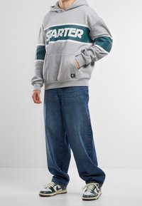 Sudadera gris con el texto "STARTER" en blanco y acentos de rayas en azul verdoso, combinada con jeans de corte holgado en azul y zapatillas verdes y blancas.