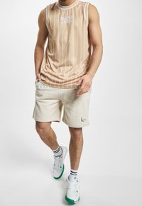 Beżowy bezrękawnik z pionowymi białymi paskami, zestawiony z jasnobeżowymi szortami. Posiada akcent logo na szortach i białe sneakersy.