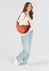 Borsa a tracolla in pelle arancione trapuntata con tracolla intrecciata e accento metallico. Il modello indossa un maglione chiaro e jeans per un contrasto.