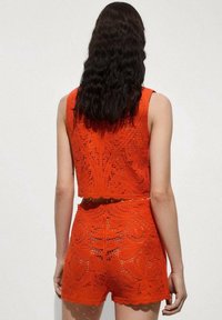 Zestaw robót na szydełku w jaskrawej pomarańczy, składający się z bezrękawowej topu i szortów z wysokim stanem. Skomplikowany wzór oraz falbaniasty dół.