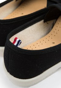 Des chaussures slip-on en toile noires avec une semelle intérieure beige, une semelle extérieure en caoutchouc blanc et une petite étiquette rouge, blanche et bleue à l'arrière.