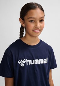 Tricou din bumbac de culoare bleu marin, cu guler rotund și mâneci scurte, având un logo grafic mare cu text alb, "hummel," pe față.
