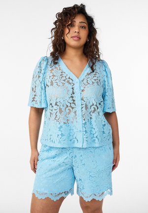 Frau trägt hellblaues, florales Spitzenoberteil mit Knöpfen, kurzen Ärmeln und passenden Spitzen-Shorts, steht vor weißem Hintergrund.