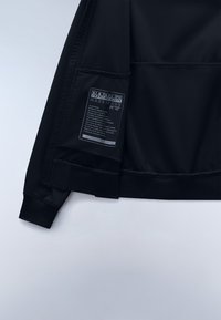 Sort jakke med netstruktur, ribbede manchetter og en label med vaskeanvisninger og materialeoplysninger på indersiden.