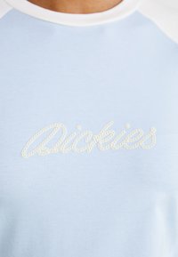 Světle modré tričko s bílým kulatým výstřihem a vyšitým logem "Dickies" ve provázkovém designu napříč hrudníkem. Hladká textura látky.