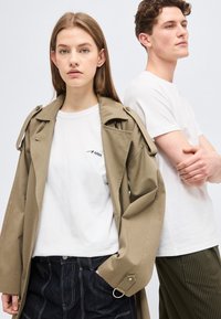 Νεαρή γυναίκα με μπεζ καπαρντίνα και λευκό t-shirt ARKK στέκεται δίπλα σε νεαρό άνδρα με λευκό t-shirt και ριγέ παντελόνι, μπροστά σε απλό φόντο.