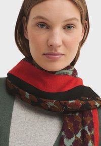 Szydełkowany szalik w ciepłych odcieniach czerwieni i czerni, z wzorzystymi akcentami w ziemistych tonacjach; miękka tekstura, owinięty wokół szyi.