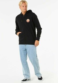 Sort lynlås hoodie med en cirkulær farverig patch på brystet; sat sammen med lyseblå baggy jeans og sorte sneakers.