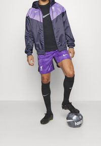 Nike tracksuit featuring a navy and purple jacket, black shirt, purple shorts with vertical stripes, black socks, and black cleats, standing on a football.