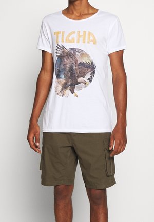 Biała koszulka z bawełny z graficznym motywem orła w okrągłym wzorze oraz napisem "TIGHA" w kolorze złotym. Połączona z oliwkowymi szortami cargo.