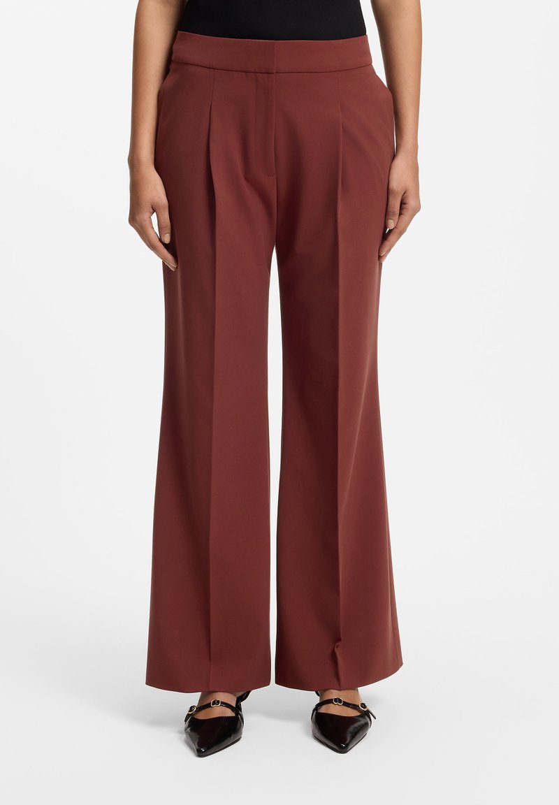 Femme portant un pantalon marron taille haute à jambes larges, associé à des ballerines noires à bout pointu sur un fond blanc uni.
