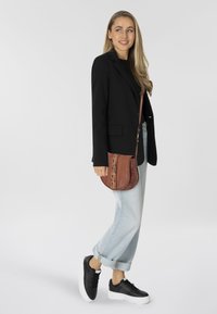 Donna in piedi e sorridente, indossa un blazer nero, jeans chiari arrotolati, sneakers nere e porta una borsa a tracolla marrone con dettagli decorativi.