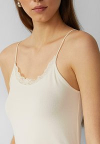 Lichtbeige cami met dunne verstelbare bandjes, kant langs de halslijn en een aansluitende textuur. Glad materiaal zonder patronen.