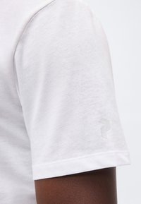 Λευκό κοντομάνικο T-shirt σε άτομο με σκούρο δέρμα, που δείχνει ένα μικρό, διακριτικό ανακλαστικό λογότυπο στο μανίκι.