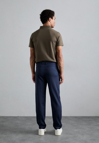 Olivovo zelená polokošeľa s krátkymi rukávmi, skombinovaná s námorníckymi nohavicami. Model stojí chrbtom k divákovi a má na sebe biele topánky.