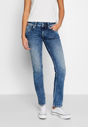 Jeans de mezclilla azul con un ajuste slim, que cuentan con cinco bolsillos, áreas deslavadas y costuras de contraste. Combinados con zapatillas blancas.