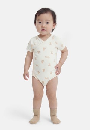 Μικρό παιδί που στέκεται, φορώντας κρεμ φορμάκι με ανοιχτά καφέ ζωάκια και μπεζ κάλτσες, με φόντο απλό λευκό τοίχο.